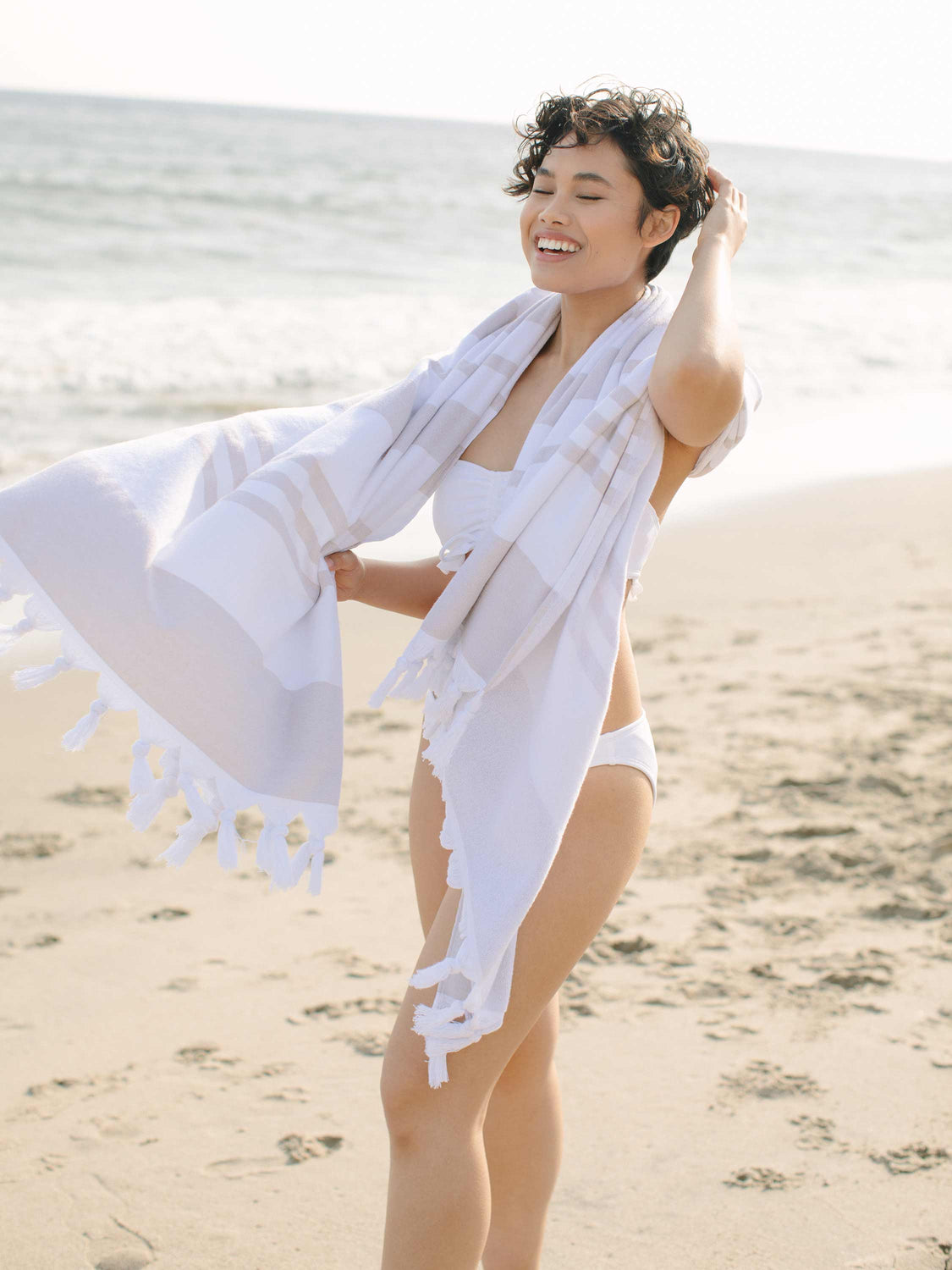 A woman standing on the beach wrapped in an oversized tan and white striped Turkish towel.