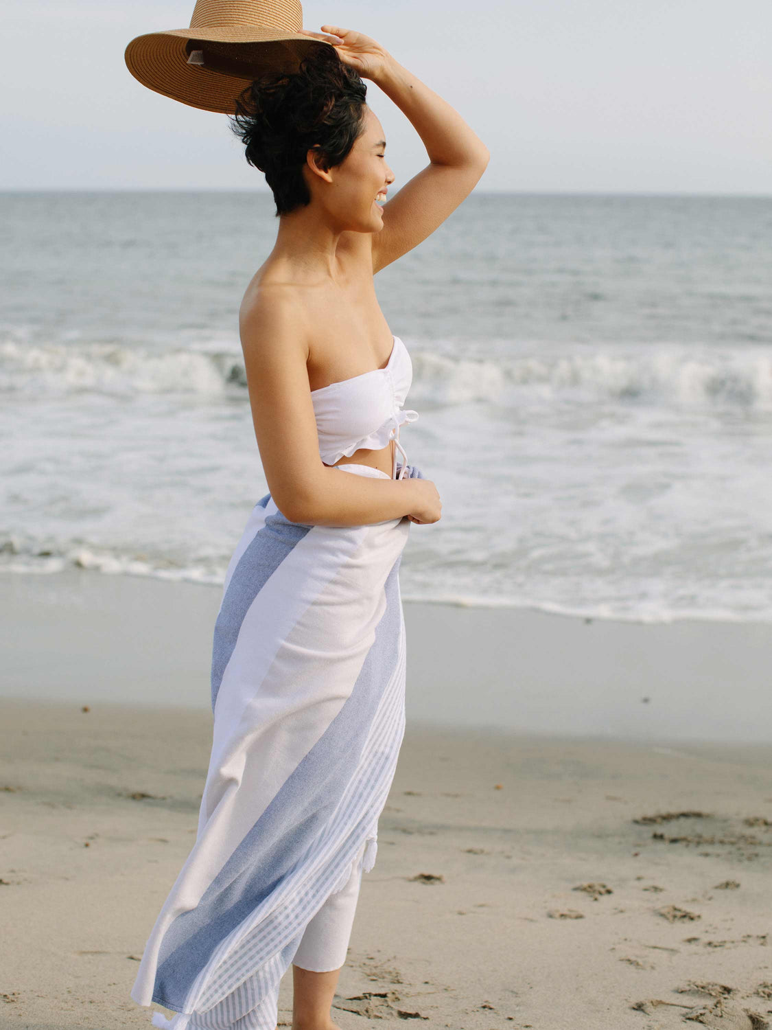 A woman standing on the beach wearing a sea and dune striped Turkish towel as a sarong.