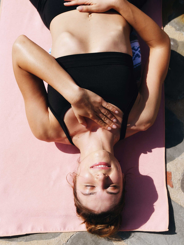A woman in black yoga clothes laying on a blush colored yoga mat towel.