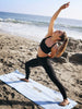 A woman on the beach in a yoga pose standing on her powder blue yoga mat towel.